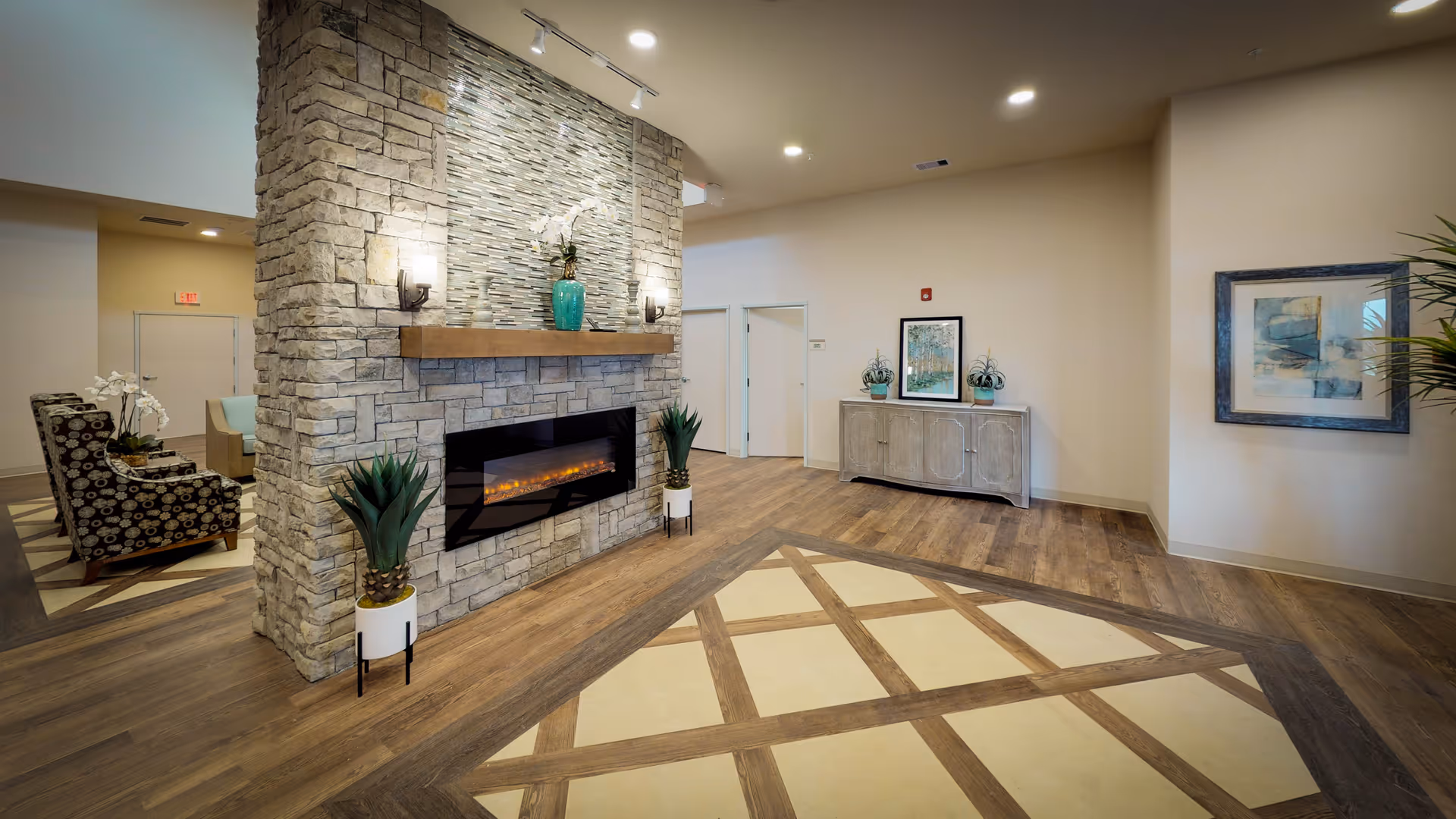 A spacious and well-lit interior common area featuring a stone fireplace with a modern electric insert. The fireplace wall is decorated with a mosaic tile design and two wall sconces. There are two potted plants on either side of the fireplace. The floor has a decorative pattern with wood and tile. In the background, there is a wooden sideboard with two plants and a framed picture above it, and another framed artwork on the adjacent wall. To the left, there is a seating area with patterned armchairs and a small table with a white orchid.