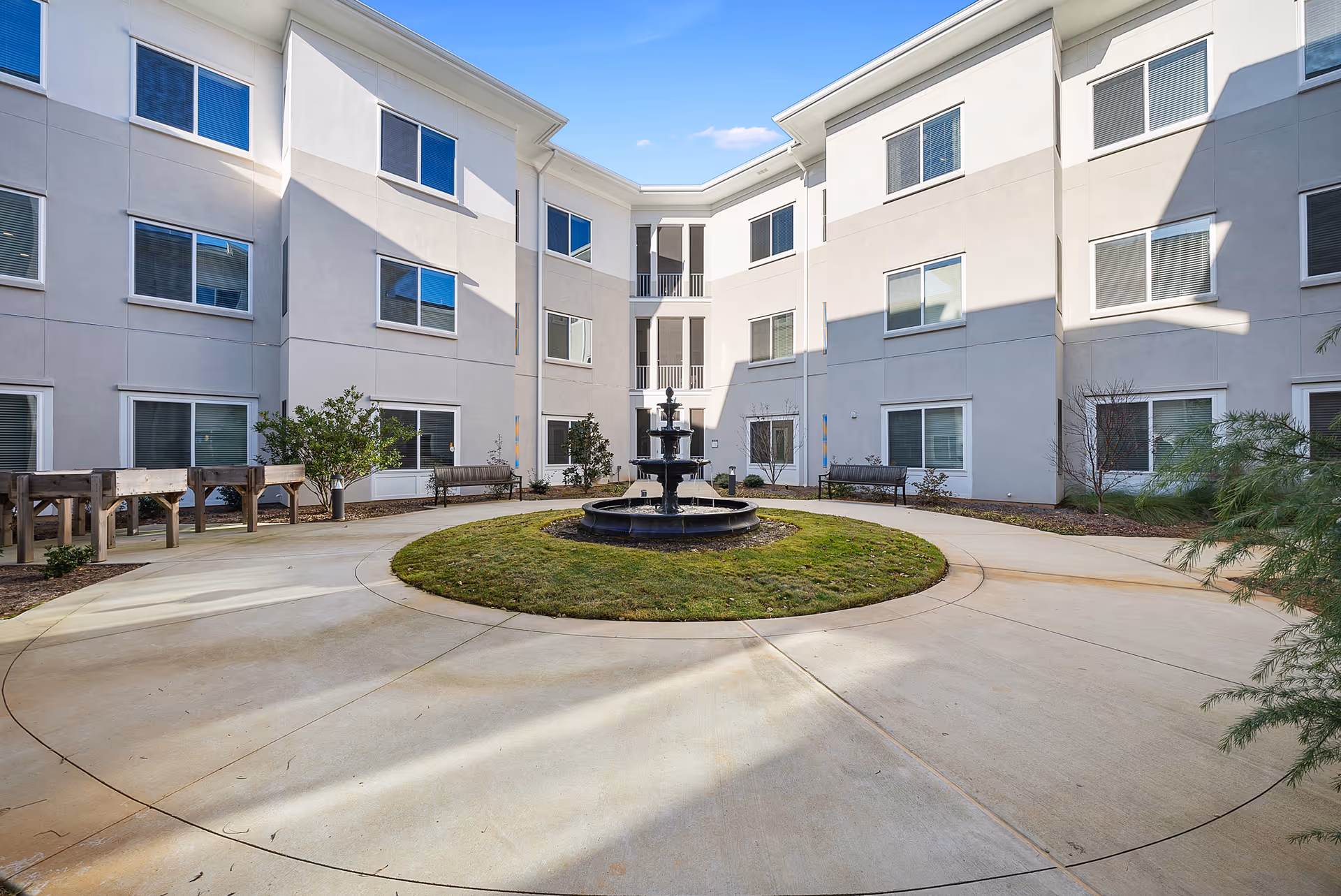 Outdoor courtyard area of a senior living facility with a circular paved walkway surrounding a grassy area with a multi-tiered water fountain in the center. The courtyard is enclosed by a three-story building with multiple windows and benches along the perimeter.