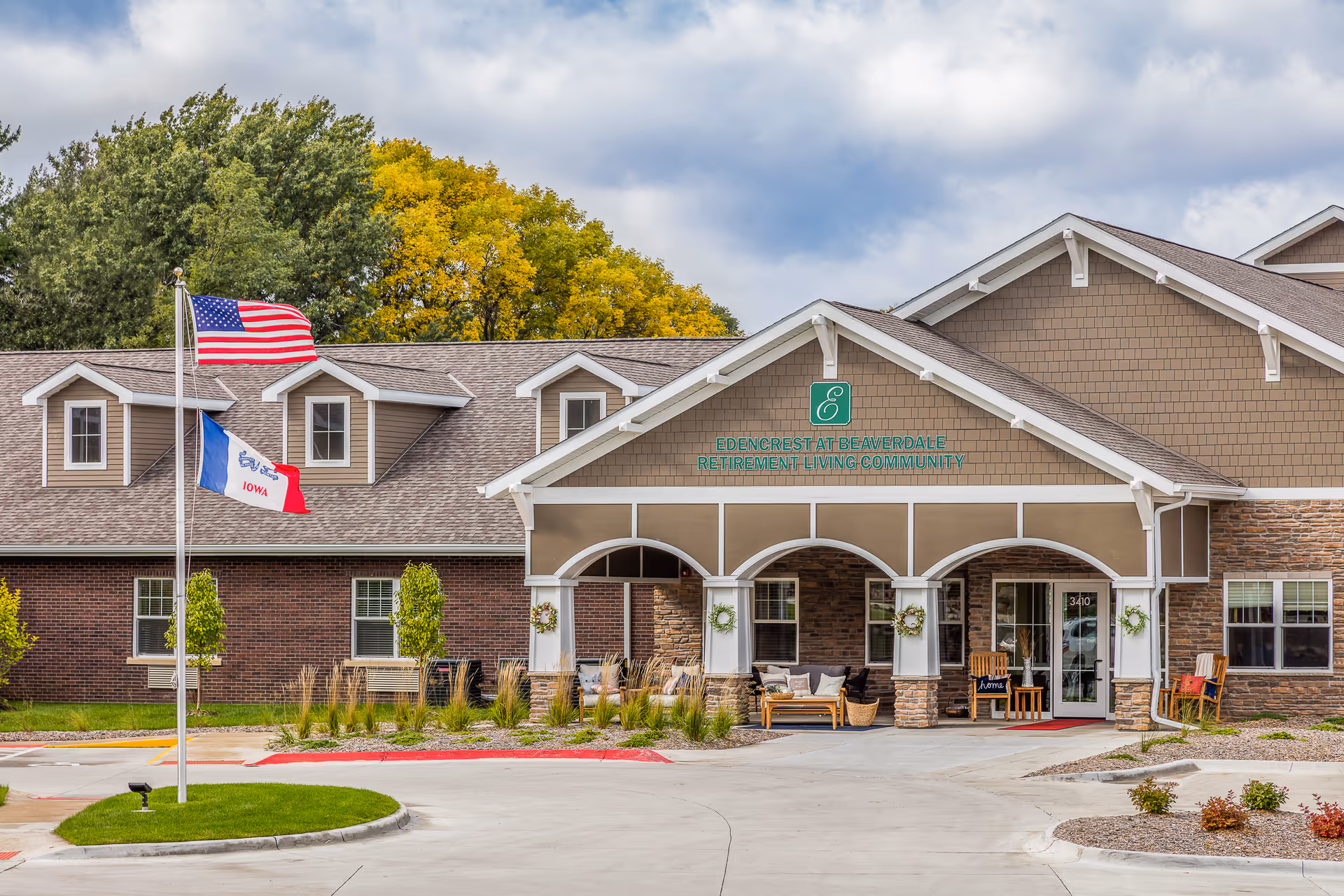 Front exterior view of Edencrest at Beaverdale retirement living community building with a covered entrance, seating area, American and Iowa state flags, and landscaped surroundings under a partly cloudy sky.