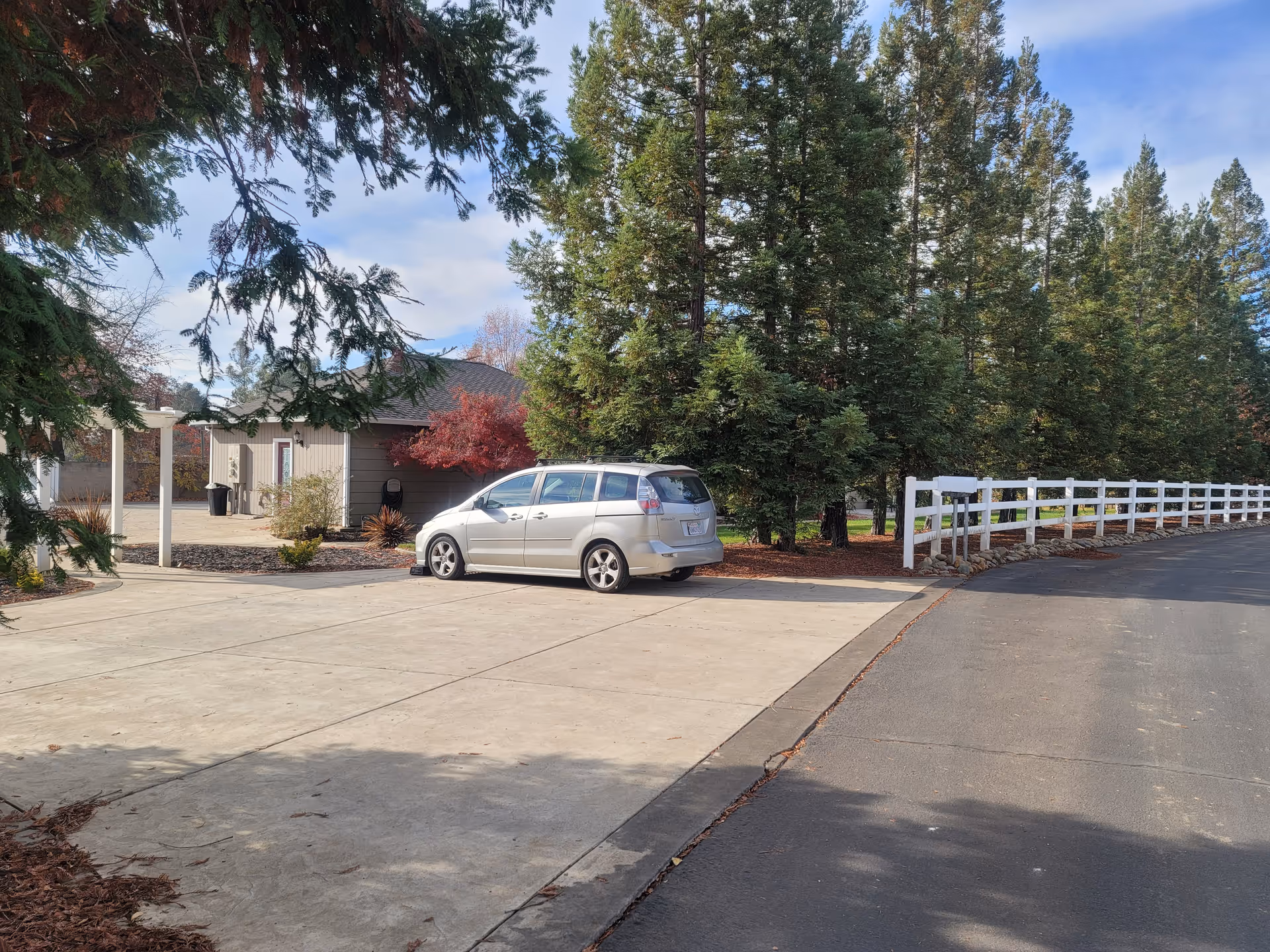 Driveway and front yard with a silver minivan parked near a small building, white fence, and tall evergreen trees.