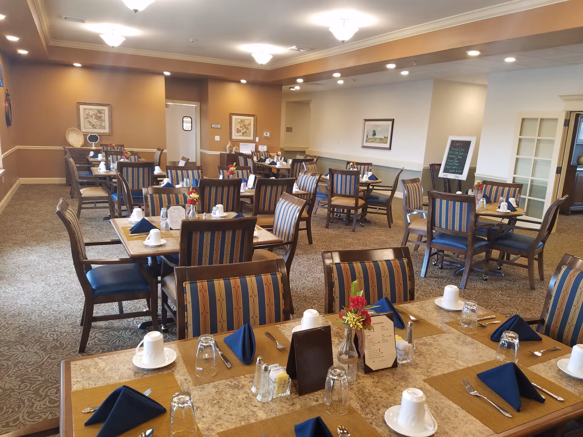 A dining room in a senior living facility with multiple tables set for meals. Each table has chairs with striped upholstery, place settings including cups, glasses, silverware, and folded navy blue napkins. There are small flower arrangements and menus on the tables. The room has warm brown and beige walls, framed artwork, and carpeted flooring.