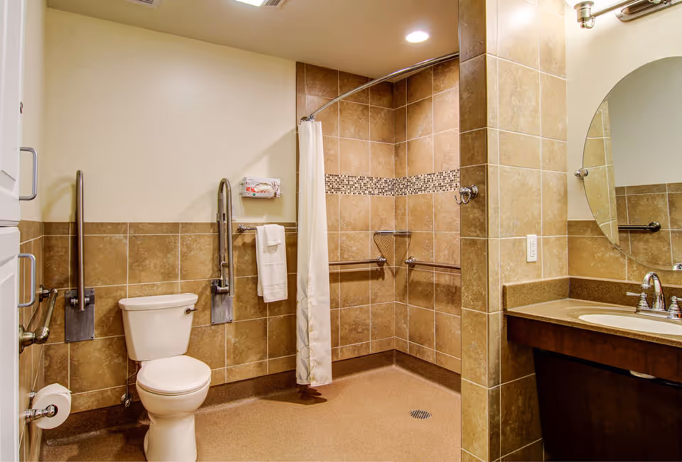 A spacious, accessible bathroom with beige tiled walls and floor. The bathroom features a white toilet with metal grab bars on both sides, a walk-in shower area with a curved shower curtain rod and grab bars, a towel rack with a white towel, and a vanity with a sink and a round mirror above it. The lighting is bright with recessed ceiling lights.