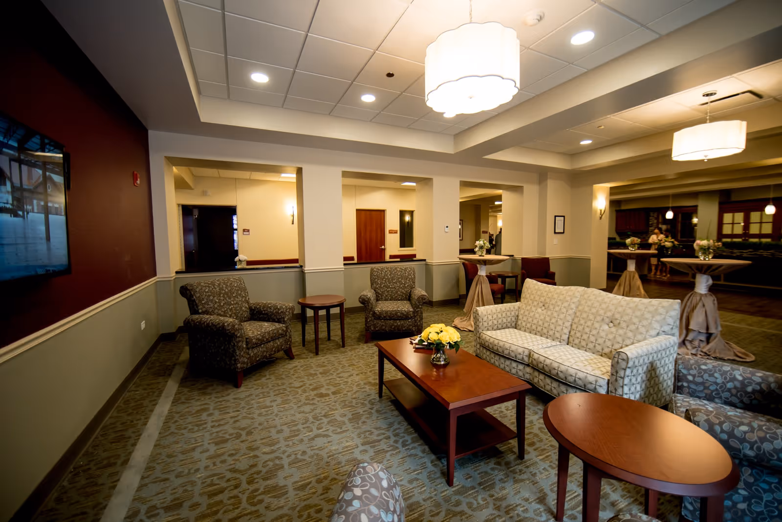 A well-lit common area with patterned armchairs, a patterned sofa, wooden coffee tables, and a large ceiling light fixture. The room has beige walls with a maroon accent wall featuring a mounted flat-screen TV. There are small round tables with floral arrangements and a carpeted floor with a subtle pattern.