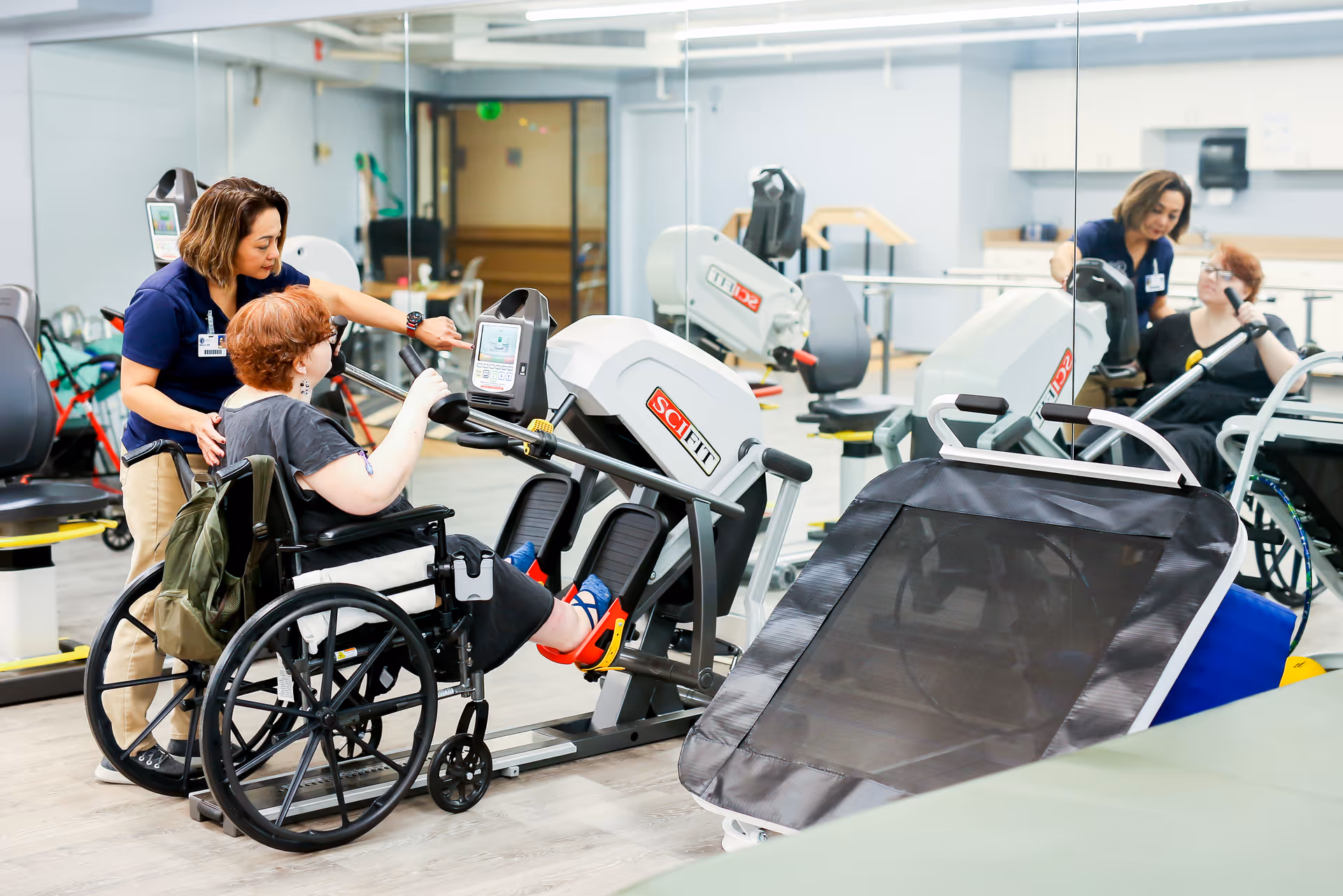 A woman in a wheelchair is exercising her legs on a SciFit exercise machine while a healthcare worker assists and encourages her in a rehabilitation center gym with mirrored walls and various exercise equipment.