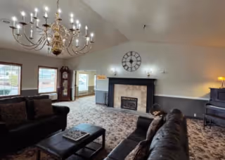 Spacious senior living lounge with sofas, a large chandelier, fireplace, grandfather clock, piano, and carpeted floor.