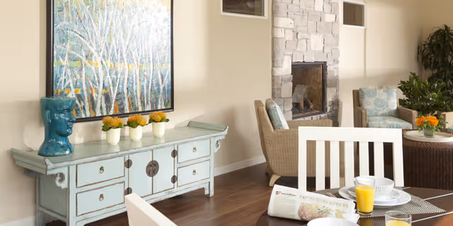 A cozy living area with a stone fireplace, two wicker armchairs with cushions, a small round table with a flower arrangement, and a light blue vintage sideboard with decorative items including a blue ceramic head and small potted plants. In the foreground, a dining table is set with plates, glasses of orange juice, and a newspaper.