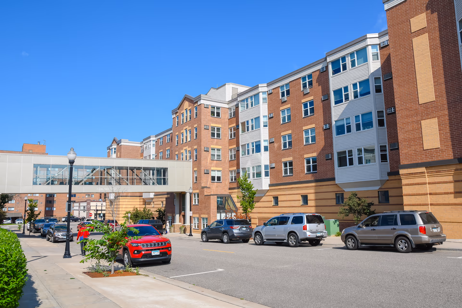 Exterior view of a multi-story brick building with several windows and a skywalk connecting to another building. Several cars are parked along the street in front of the building under a clear blue sky.