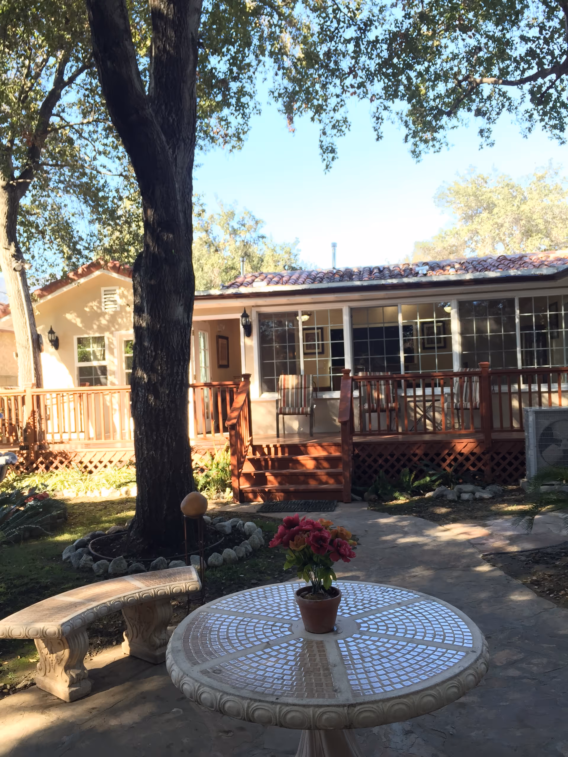 Outdoor patio with a round mosaic table holding a potted flower, a stone bench, large tree, and a raised wooden deck in front of a single-story building.