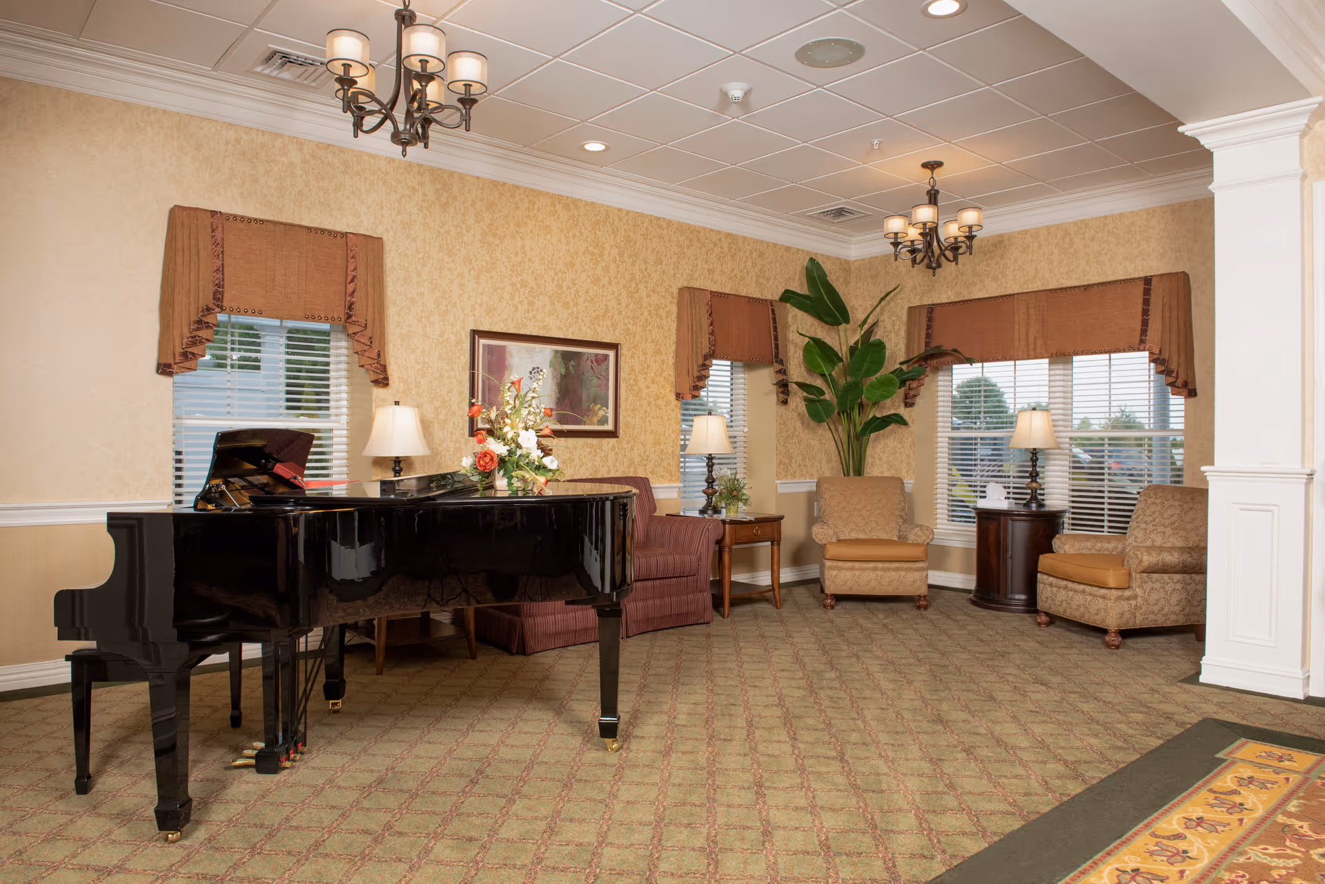 A cozy sitting area in a retirement community featuring a black grand piano with a floral arrangement on top, three upholstered armchairs, two side tables with lamps, large windows with brown valances, a potted plant, and warm beige walls with framed artwork.
