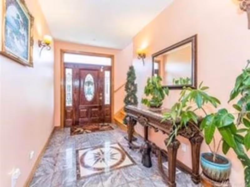 Bright entry foyer with an ornate wooden front door, decorative console table, mirror and potted plants on a patterned tiled floor.