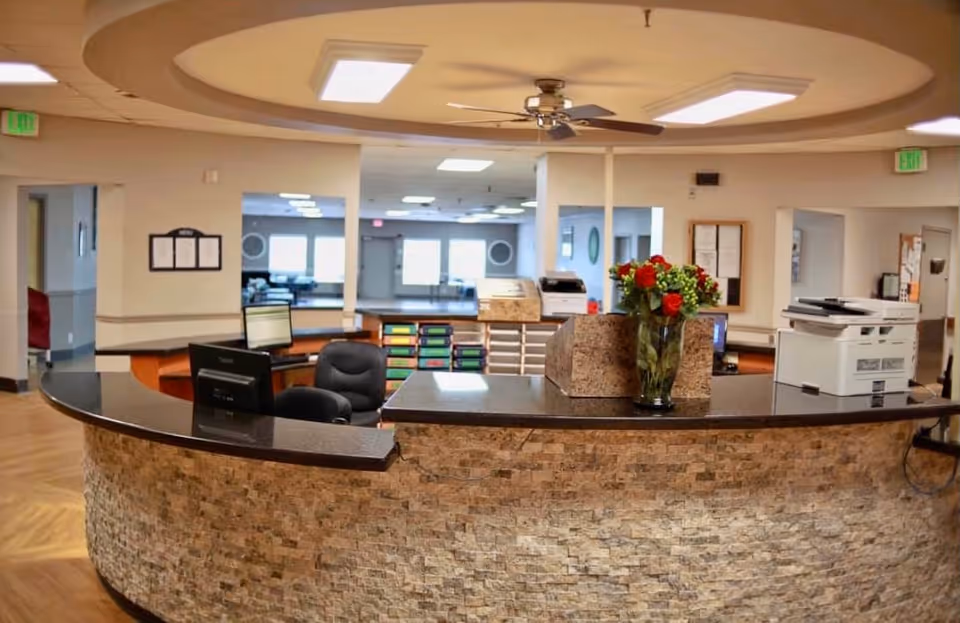 Reception desk area in a health and rehabilitation facility with a curved stone front counter, two computer monitors, office chairs, a vase with red and white flowers, and ceiling lights with a fan. The background shows open doorways and a spacious interior with natural light coming through windows.