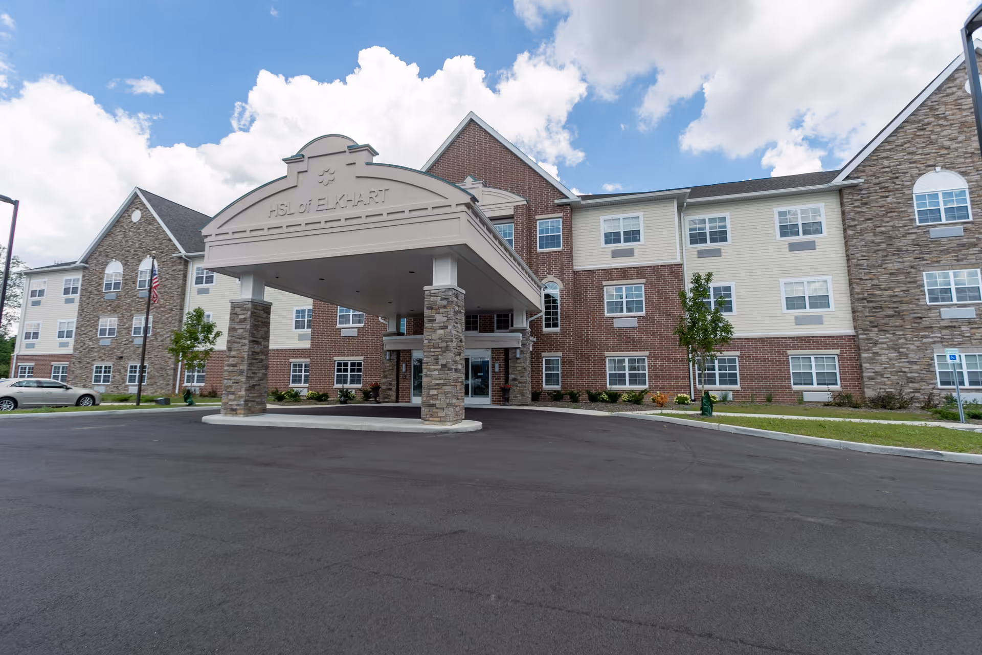 Front exterior of Hellenic Senior Living of Elkhart showing the building facade, covered entrance porte-cochere, and paved driveway under a partly cloudy sky.