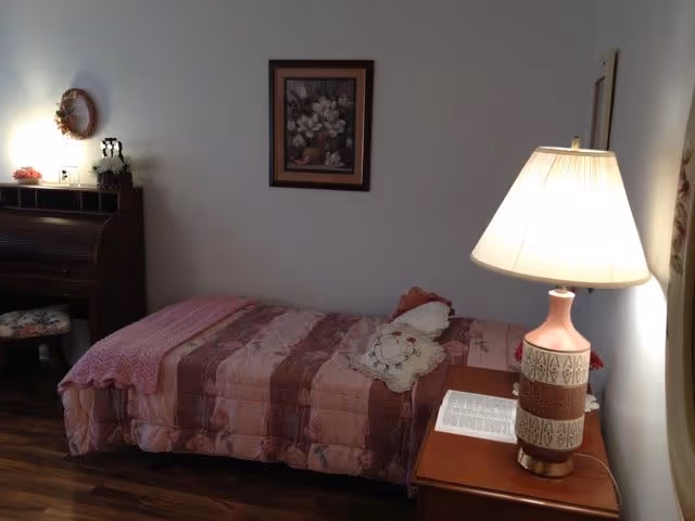 A small bedroom with a single bed covered in a pink and brown quilt. There is a decorative pillow on the bed. Next to the bed is a wooden nightstand with a lamp and an open book. On the left side of the room, there is a wooden roll-top desk with a floral cushioned stool. A framed floral picture hangs on the wall above the bed.