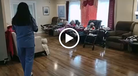 A caregiver in blue scrubs stands in a room with wooden flooring, facing three elderly individuals seated in recliners near a window with red curtains. One elderly person is using a walker, and the room is softly lit with a floor lamp and natural light from the window.