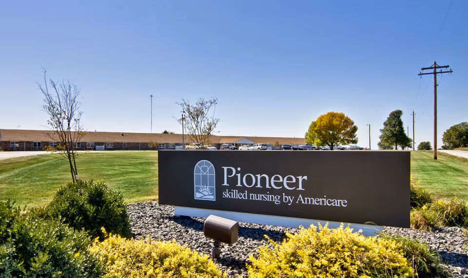 Outdoor view of the Pioneer skilled nursing facility sign with a large building and parking lot in the background under a clear blue sky. The sign is surrounded by green grass, bushes, and small trees.