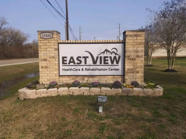 A brick sign for EastView HealthCare & Rehabilitation Center with the address number 15880 on the left pillar. The sign is surrounded by a small garden bed with decorative plants and flowers, situated on a grassy area near a road.