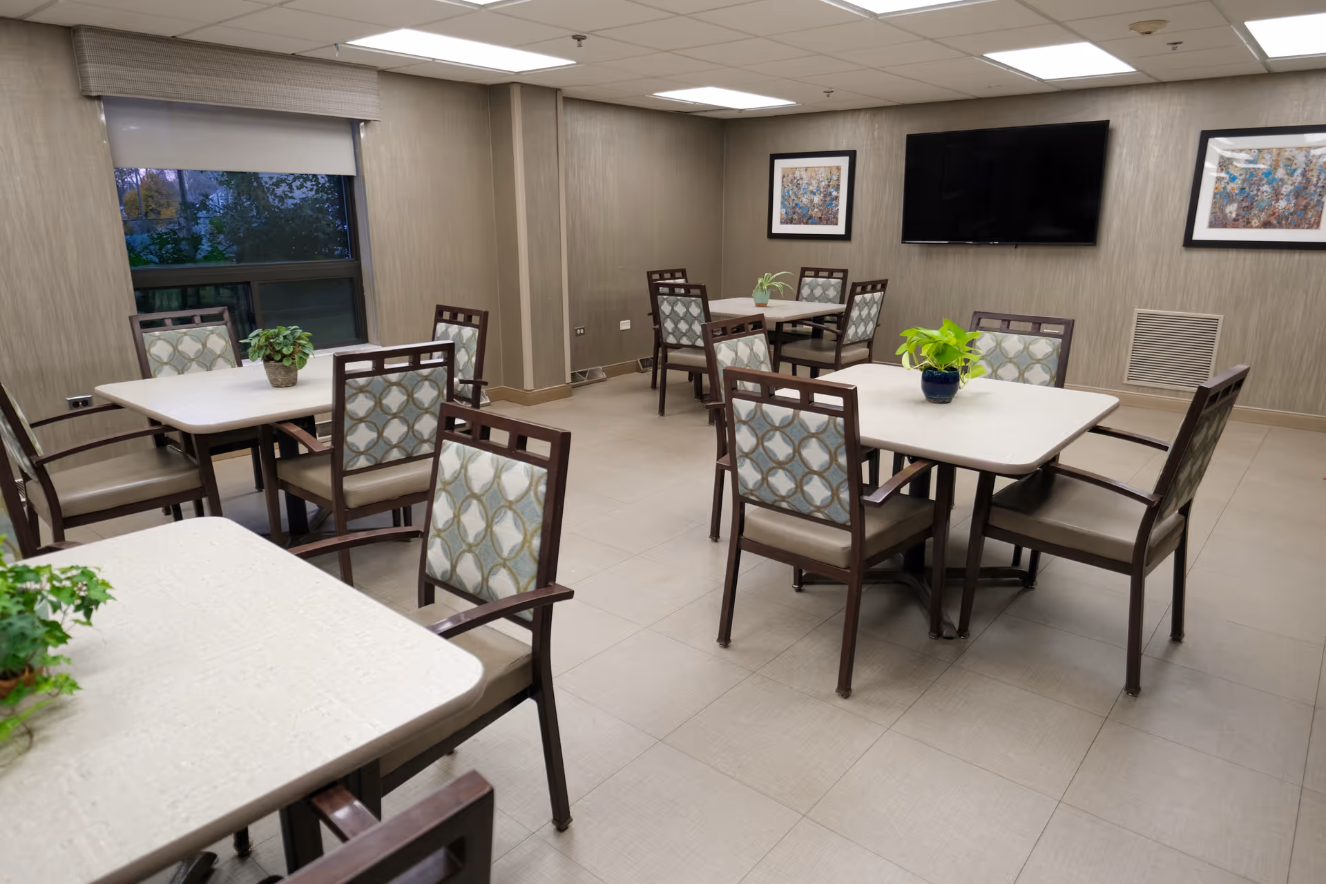 A clean communal dining room with several tables and patterned chairs, potted plants, and a wall-mounted TV.