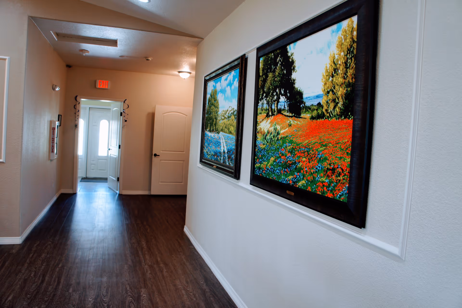A hallway with dark wood flooring and light-colored walls, featuring two framed landscape paintings on the right wall. At the end of the hallway, there is a door with glass panels letting in natural light and an exit sign above it.