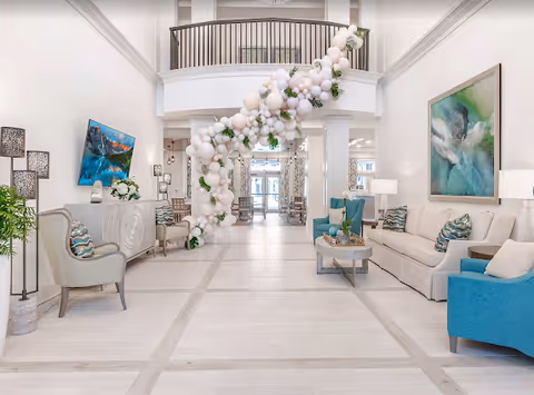 Bright and spacious living room area with white walls and light wood flooring. The room features a white sofa with blue and patterned cushions, a blue armchair, a beige armchair, and a coffee table with decorative items. A large abstract painting hangs on the wall above the sofa. A white sideboard with a TV mounted above it and decorative items is on the left side. A large balloon decoration with white and blush pink balloons and greenery extends from the floor to the second-floor balcony railing. The space is well-lit with natural light coming from the glass doors at the far end.