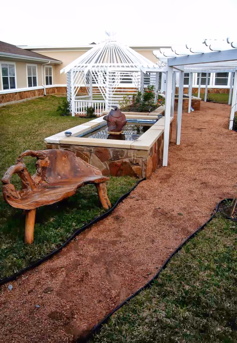 Outdoor garden area at Georgetown Living featuring a gravel pathway, a rustic wooden bench, a rectangular water feature with a decorative sculpture, white pergolas, and a white gazebo surrounded by grass and the building exterior.