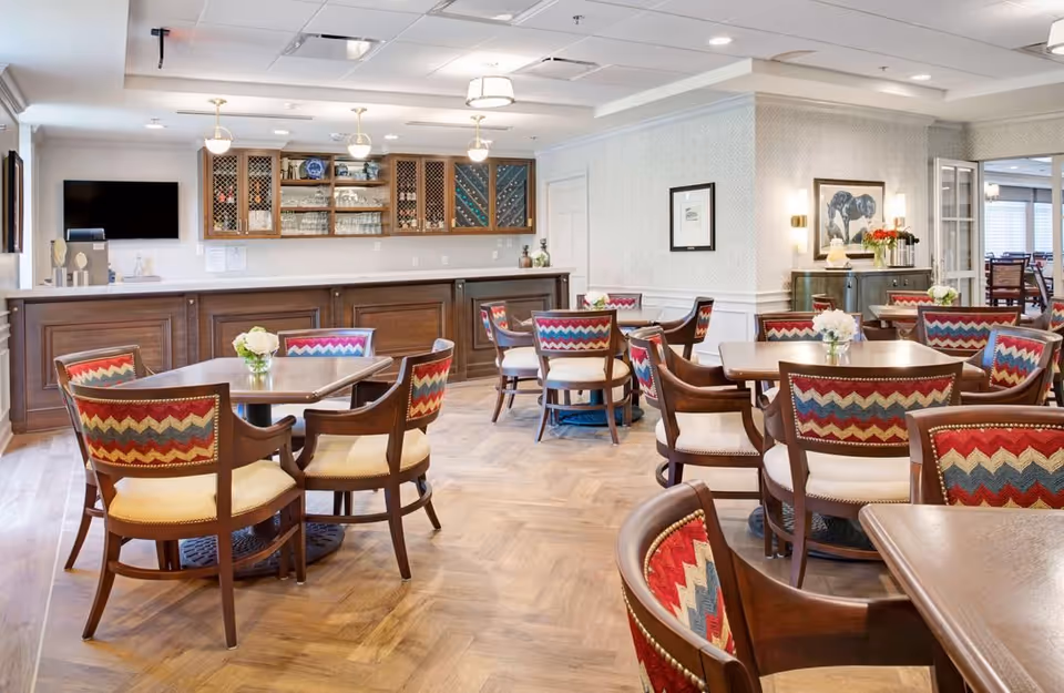Spacious dining room with round wooden tables and colorful upholstered chairs arranged before a serving counter and cabinets.