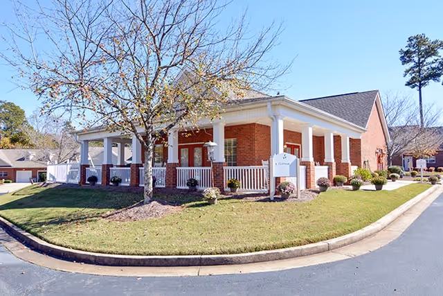 Brick single-story senior living building with a wraparound porch, white columns, a small sign near the entrance, and a landscaped lawn under a clear blue sky.