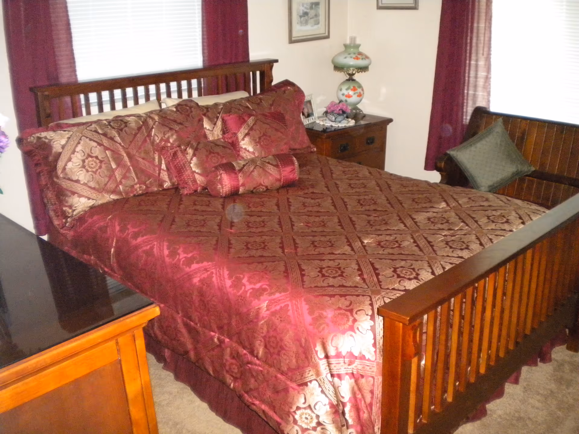 A bedroom with a wooden bed frame and a bed covered in a patterned maroon and gold bedspread with matching pillows. There is a wooden nightstand with a decorative lamp and flowers, a wooden dresser, and a wooden bench with a green cushion. The room has windows with white blinds and maroon curtains.