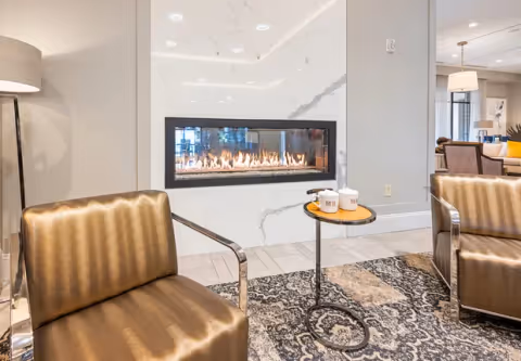 Modern lounge area with two metallic armchairs facing a linear gas fireplace set in a marble wall, a small round side table with mugs, and a patterned rug.