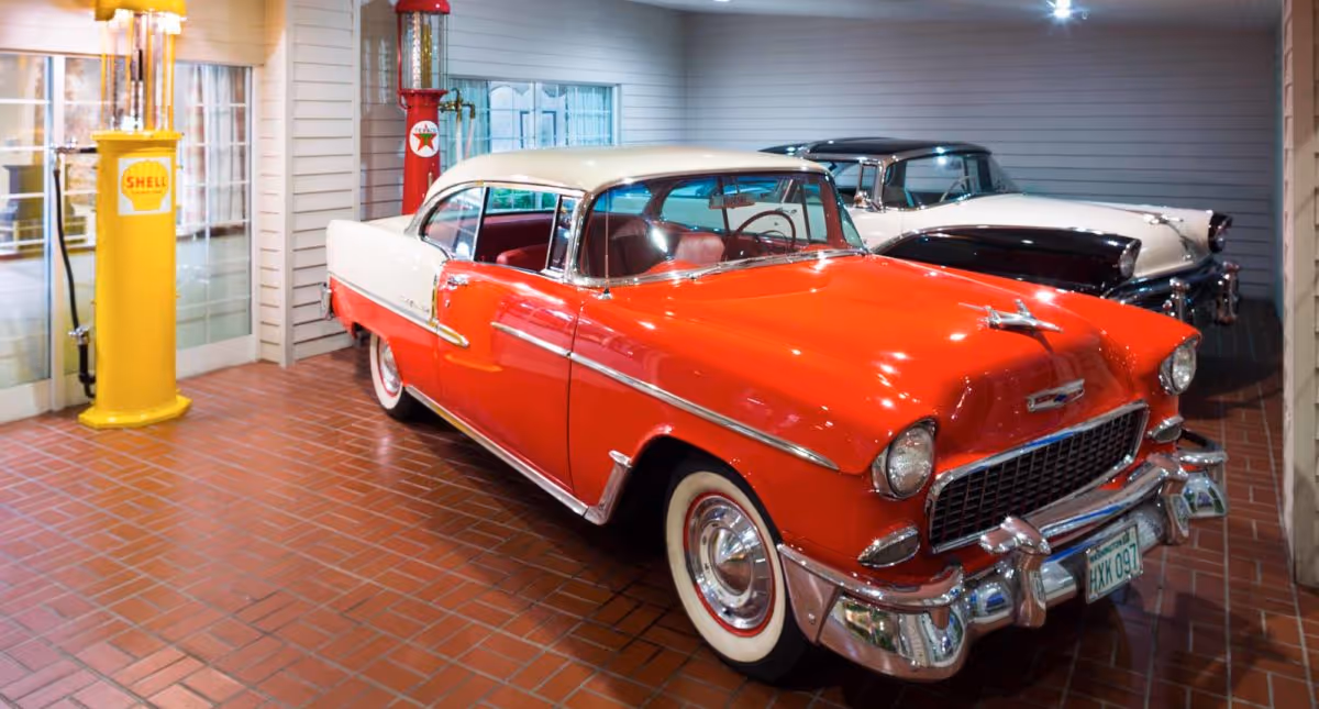 A vintage red and white classic car parked indoors on a brick floor, next to a yellow Shell gas pump and a red Texaco gas pump, with another classic car visible in the background.