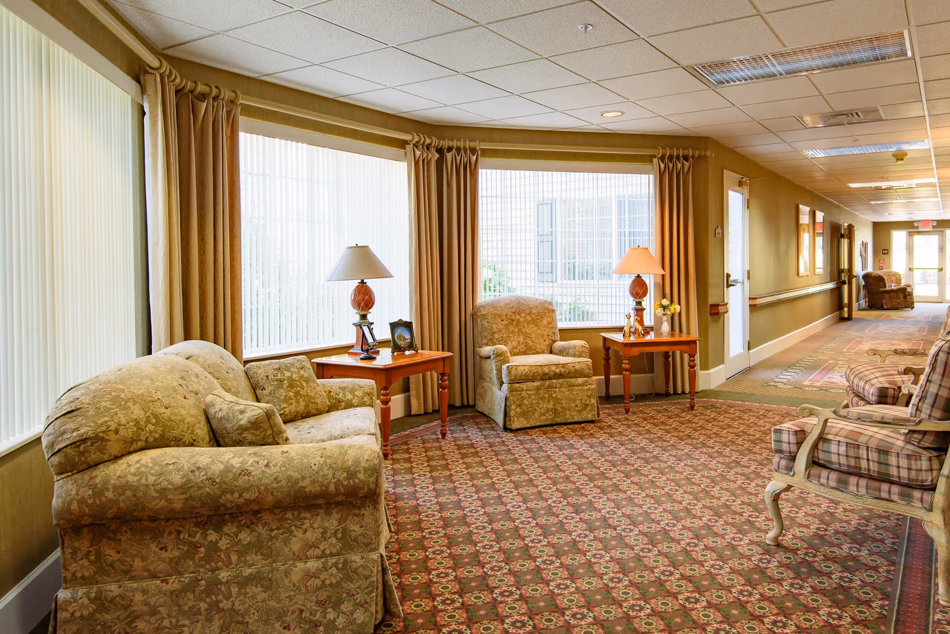 A cozy sitting area in a senior living facility hallway with floral patterned armchairs and sofa, wooden side tables with lamps and picture frames, large windows with vertical blinds and curtains, and a patterned carpet extending down the hallway.