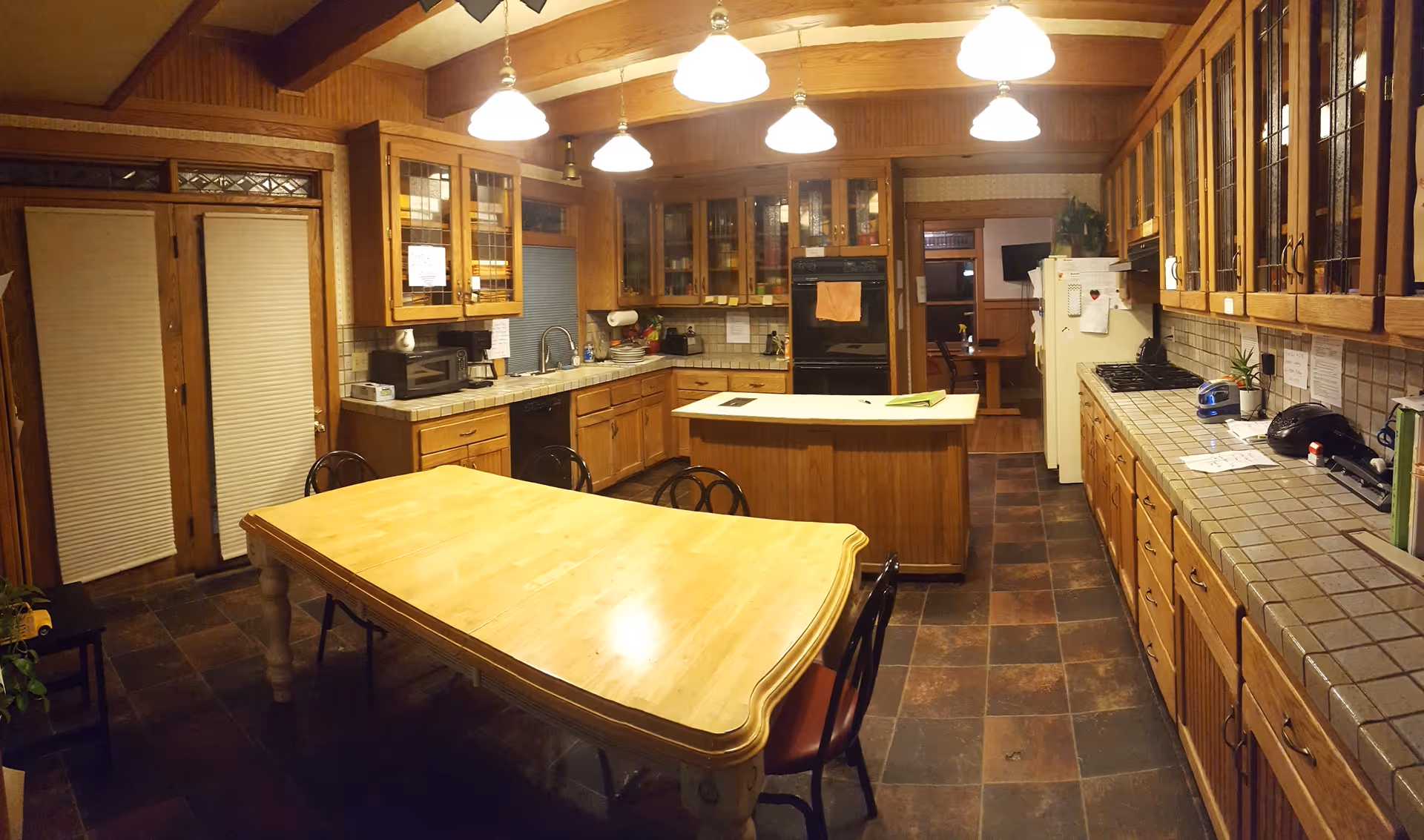 A spacious kitchen with wooden cabinets and a large wooden dining table with chairs. The kitchen features tiled countertops, a central island, a refrigerator, a built-in oven, a microwave, and overhead lighting. There are glass-front cabinets and a door with blinds on the left side.