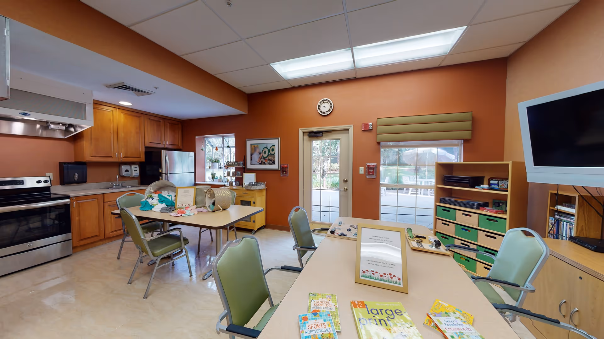 A well-lit room with a kitchen area featuring wooden cabinets, a stainless steel stove, and refrigerator. There are two tables with green chairs around them, some craft supplies and papers on the tables. A wall clock is above a door with windows on either side, showing an outdoor patio area. A flat-screen TV is mounted on the right wall above a cabinet with shelves containing books and storage bins.