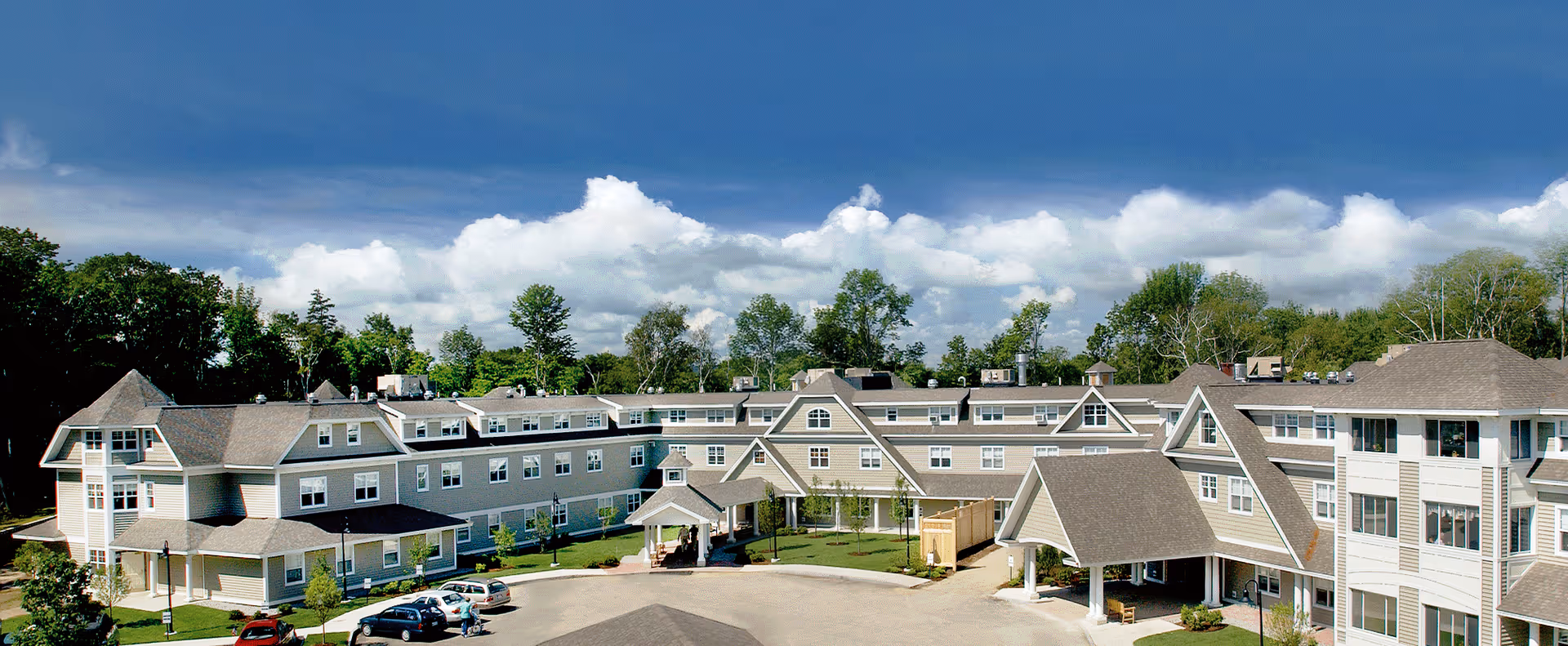 Wide exterior view of a large senior living facility building with multiple windows, peaked roofs, and a covered entrance. Several cars are parked in front, and trees and a partly cloudy sky are visible in the background.