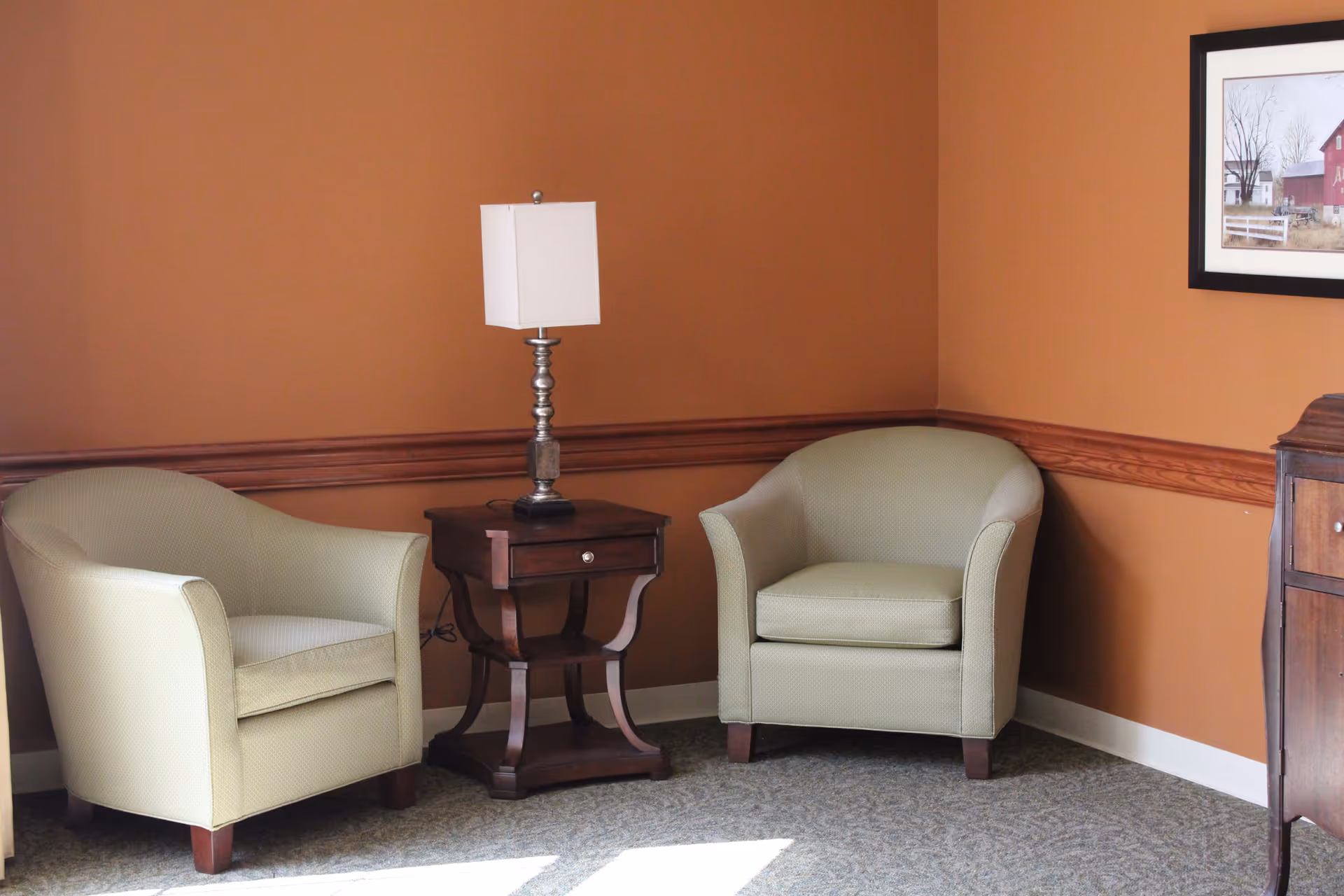 A cozy corner of a room with two light green upholstered armchairs separated by a dark wooden side table with a silver lamp on top. The walls are painted a warm brown color with a wooden chair rail. A framed picture hangs on the wall above a wooden cabinet on the right side.