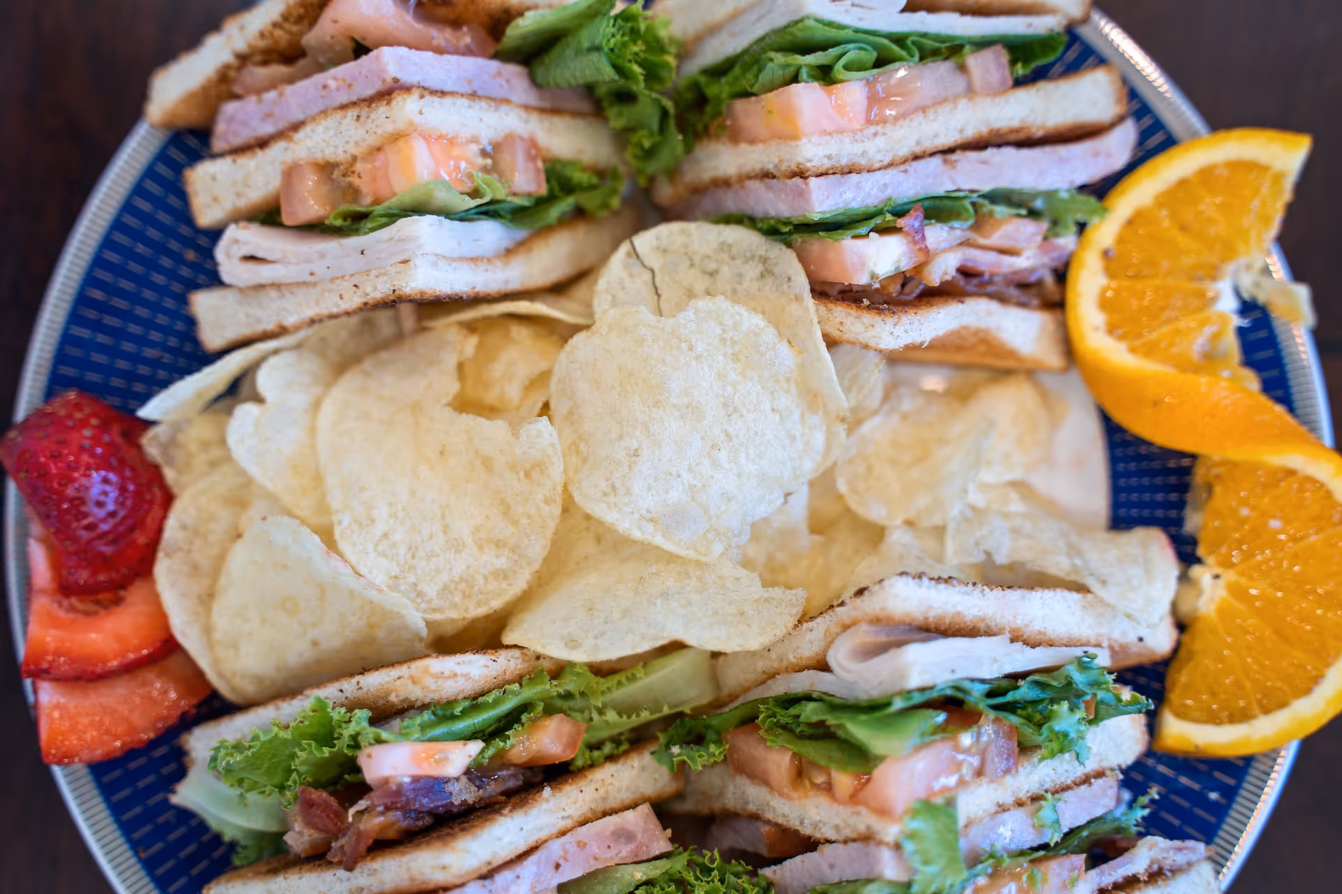 A plate with club sandwiches containing lettuce, tomato, turkey, and bacon, served with potato chips, sliced strawberries, and orange wedges.