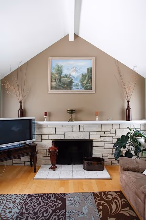 A cozy living room with a stone fireplace centered on a beige wall. Above the fireplace hangs a framed landscape painting depicting mountains, trees, and a waterfall. On the mantel are decorative items including two tall vases with dried branches, two candles, and a small ornamental bowl. To the left of the fireplace is a wooden table with a flat-screen TV, and to the right is a brown upholstered chair. The floor is wooden with a patterned area rug in front of the fireplace.