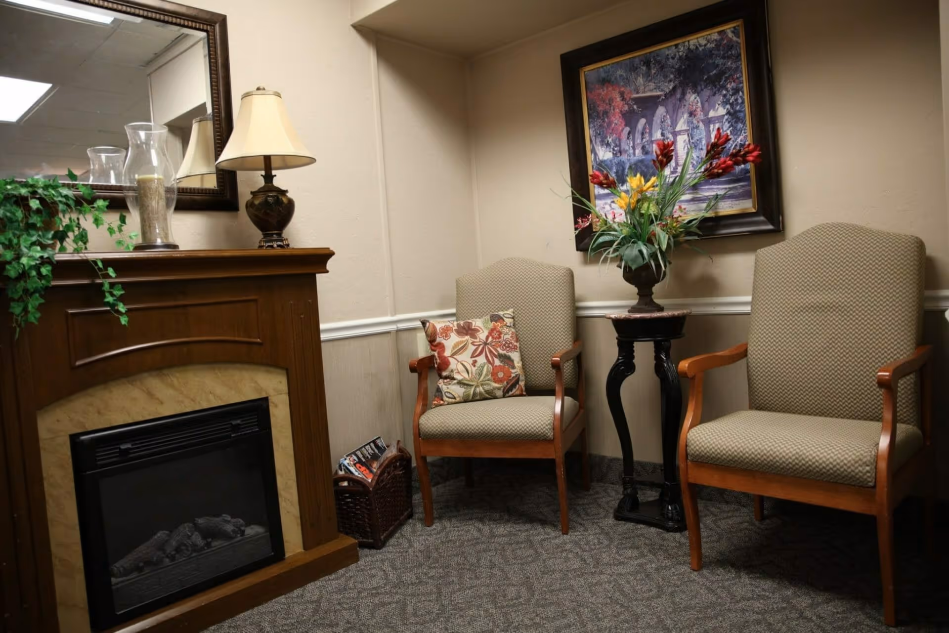 A cozy seating area with two upholstered chairs, a small side table holding a floral arrangement, and a fireplace mantel with a lamp and mirror.