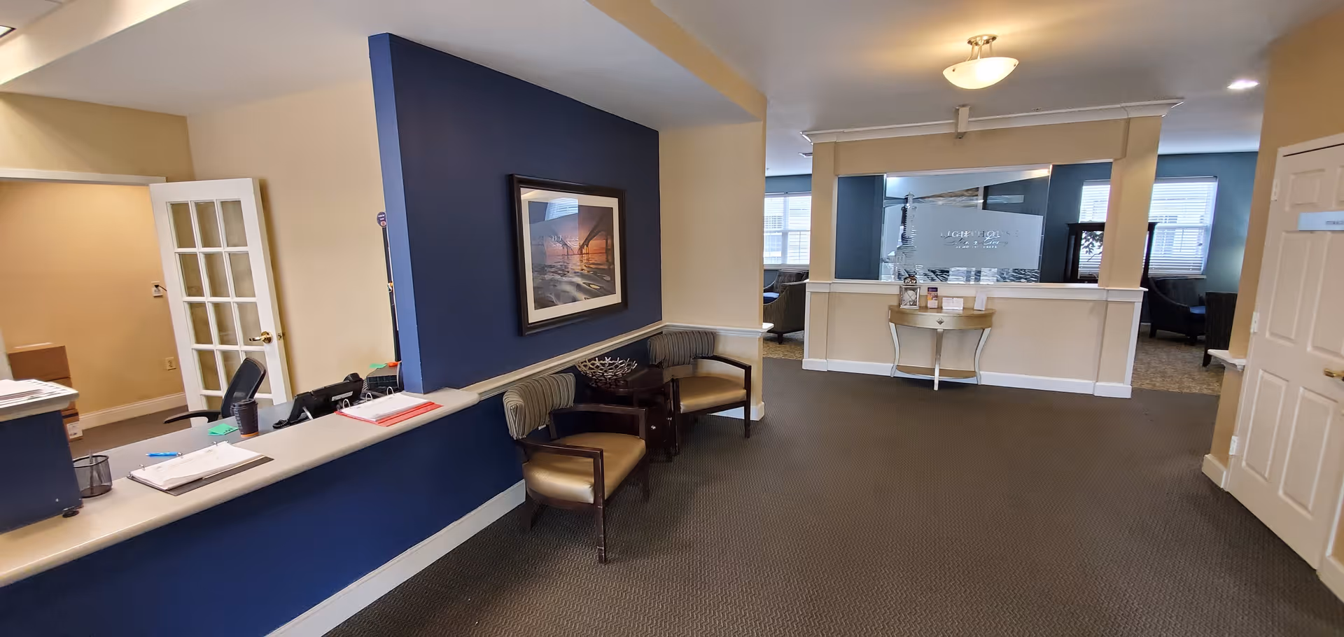Reception area of a senior living facility with a blue and beige color scheme. There is a reception desk on the left with office supplies and a chair behind it. Two chairs and a small table with decorative items are placed against a blue accent wall with a framed picture. In the background, there is a hallway leading to a common area with windows and additional seating.