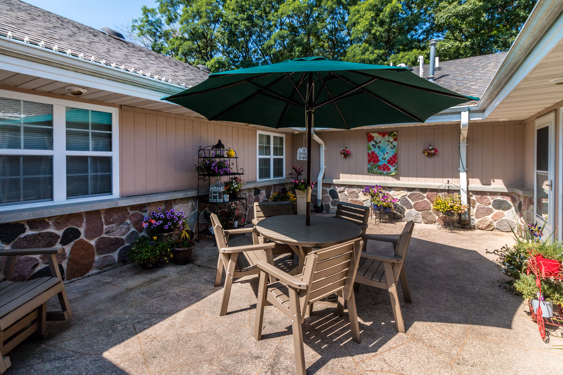Outdoor patio area with a round table and four chairs under a large green umbrella. The patio is surrounded by a building with stone and wood siding, decorated with various potted plants and flowers. There is a decorative wall hanging and a metal plant stand with more flowers. Trees and blue sky are visible in the background.