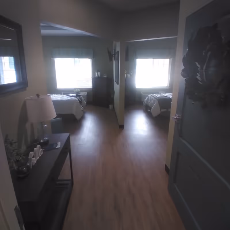 View down a hallway in a senior living facility showing two bedrooms on either side, each with a bed, window with blinds, and a dresser. The hallway has wood flooring and a small table with a lamp and decorative items on the left side. A wreath hangs on the door to the right.