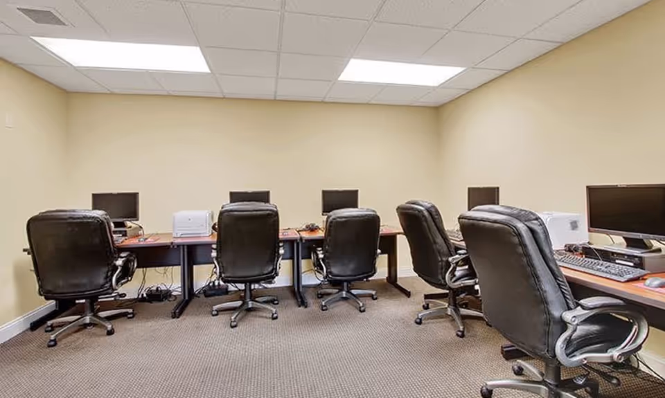 A computer room with six black office chairs placed in front of desks that hold desktop computers and a printer. The room has beige walls, a carpeted floor, and a drop ceiling with fluorescent lighting.