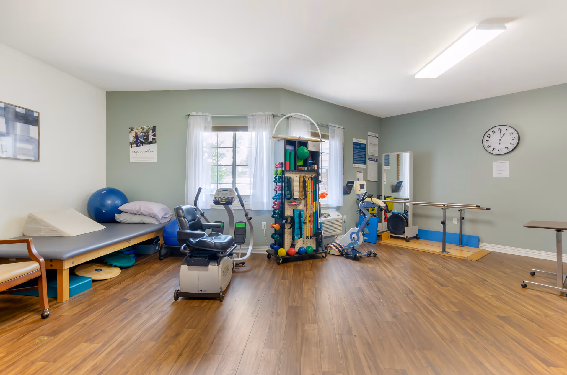 A physical therapy room with exercise equipment including a recumbent bike, parallel bars, therapy balls, and a treatment table with pillows. The room has wood flooring, light green walls, two windows with sheer white curtains, a wall clock, and various therapy tools organized on a rack.