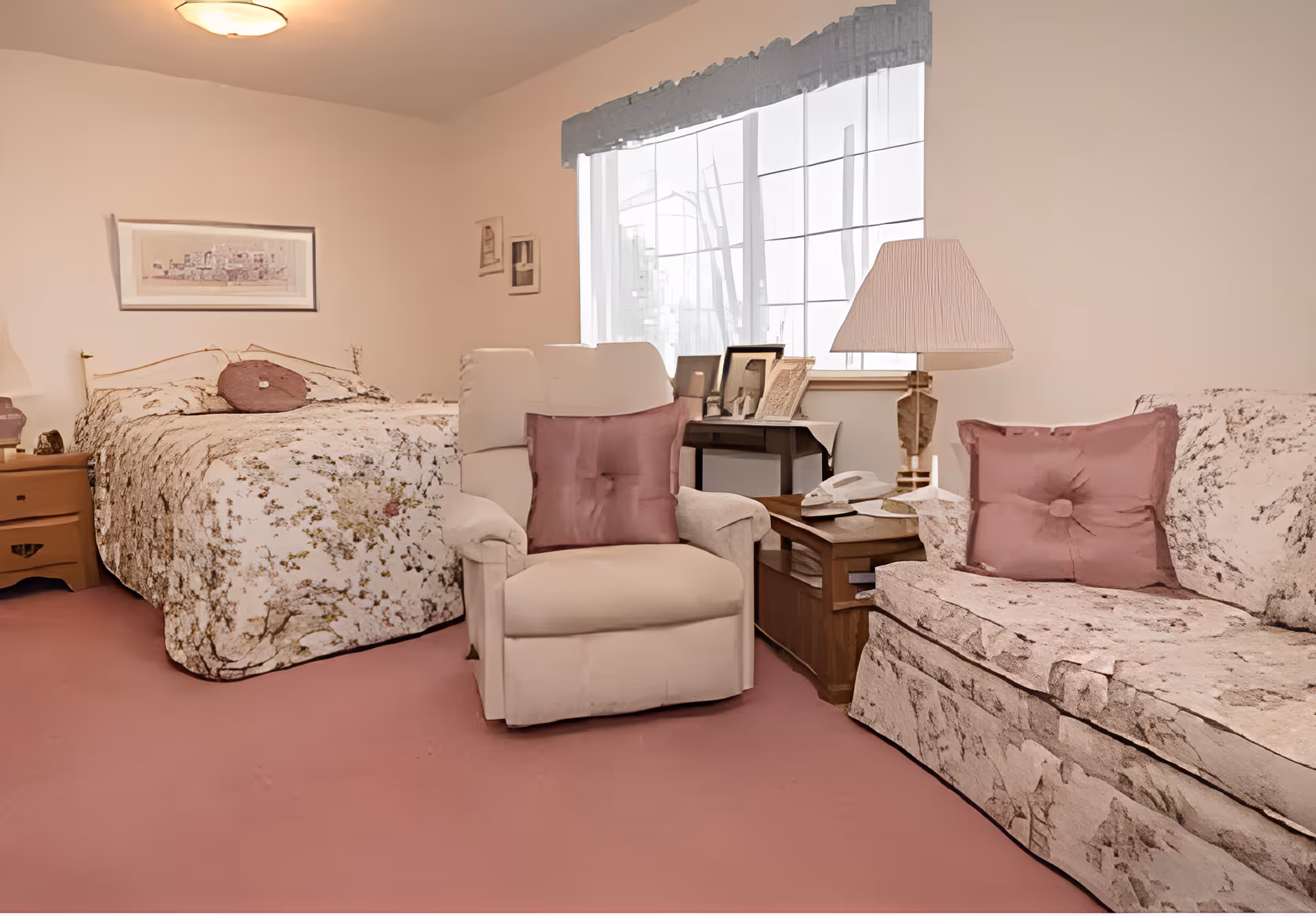 A cozy room with a bed covered in a floral bedspread, a white armchair with a mauve cushion, and a matching floral-patterned sofa with a mauve cushion. There is a wooden side table with a lamp, a telephone, and framed photos near a window with sheer curtains.