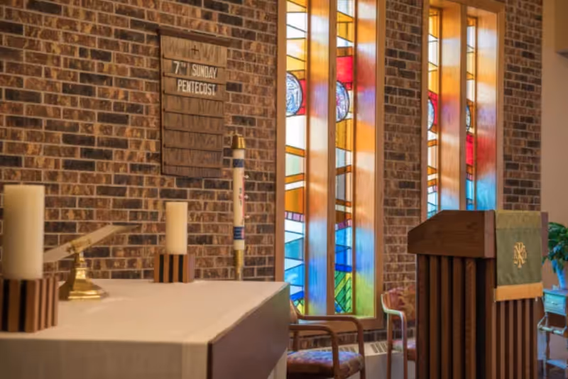 Interior view of a chapel with a brick wall, stained glass windows, an altar with candles, and a wooden lectern draped with a green cloth.