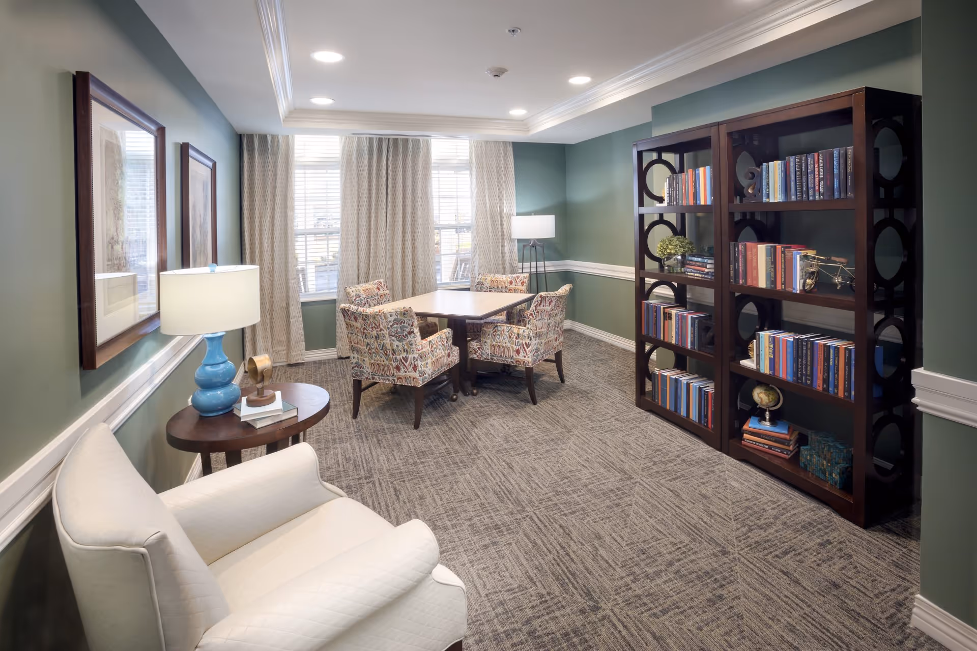 A cozy sitting room with green walls and carpeted floor featuring a white armchair, a wooden side table with a blue lamp and decorative items, a square wooden table surrounded by four patterned upholstered chairs, and two dark wooden bookshelves filled with books and decorative objects. Large windows with beige curtains allow natural light into the room.