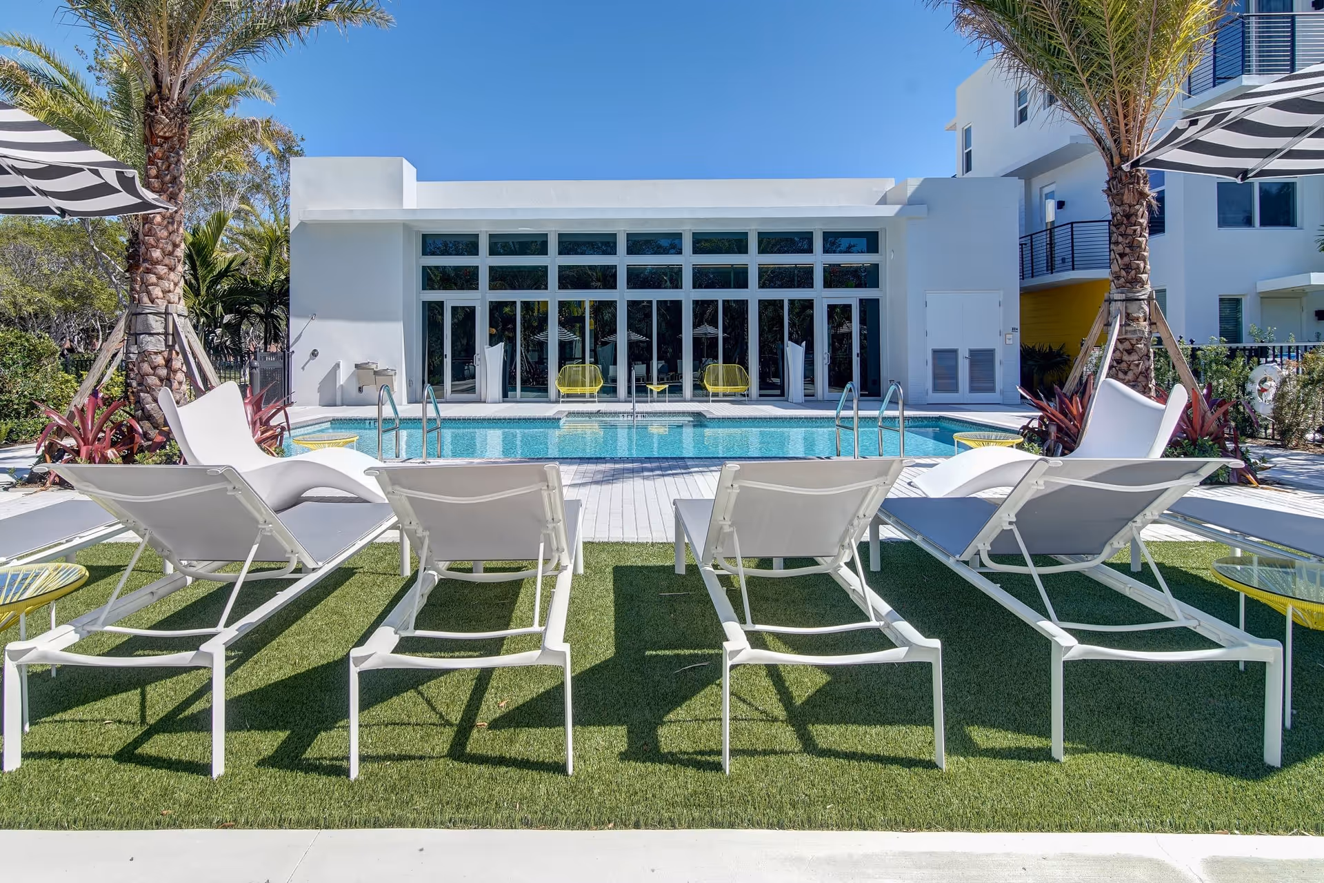 Outdoor pool area at Upside Miami featuring four white lounge chairs on artificial grass, two white modern curved chairs, yellow side tables, palm trees, and a white building with large glass windows and doors in the background under a clear blue sky.