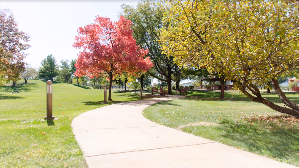A winding concrete pathway through a green grassy area with trees showing autumn colors, including a vibrant red tree and several green and yellow-leaved trees. There are benches and lamp posts along the path, and a gazebo is visible in the background under the trees.
