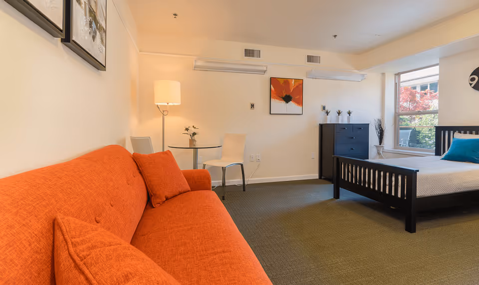 Studio-style room with an orange sofa, bed, dresser, and a small table by a floor lamp.