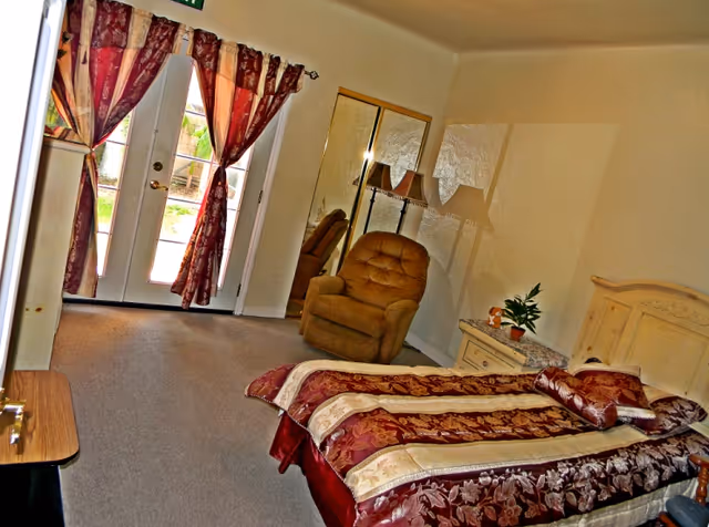 A cozy bedroom in The Villa Senior Home featuring a bed with a patterned maroon and beige bedspread, a cushioned armchair, a nightstand with a plant and a lamp, mirrored closet doors, and French doors with maroon curtains leading outside.