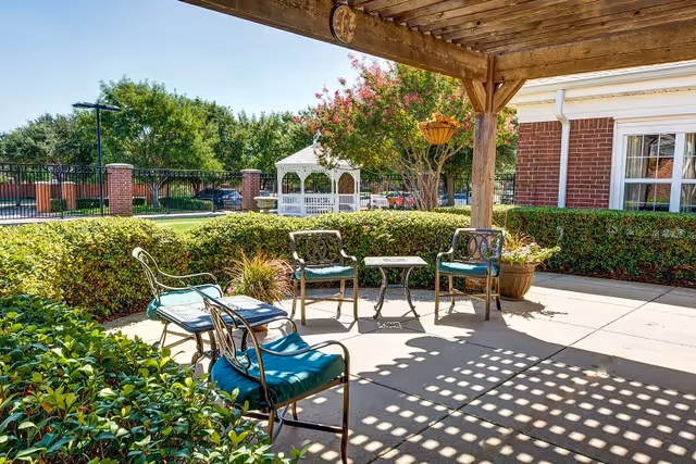 Outdoor patio area with metal chairs featuring teal cushions and a small table under a wooden pergola. Surrounding the patio are green bushes and trees, with a white gazebo and a brick building visible in the background.